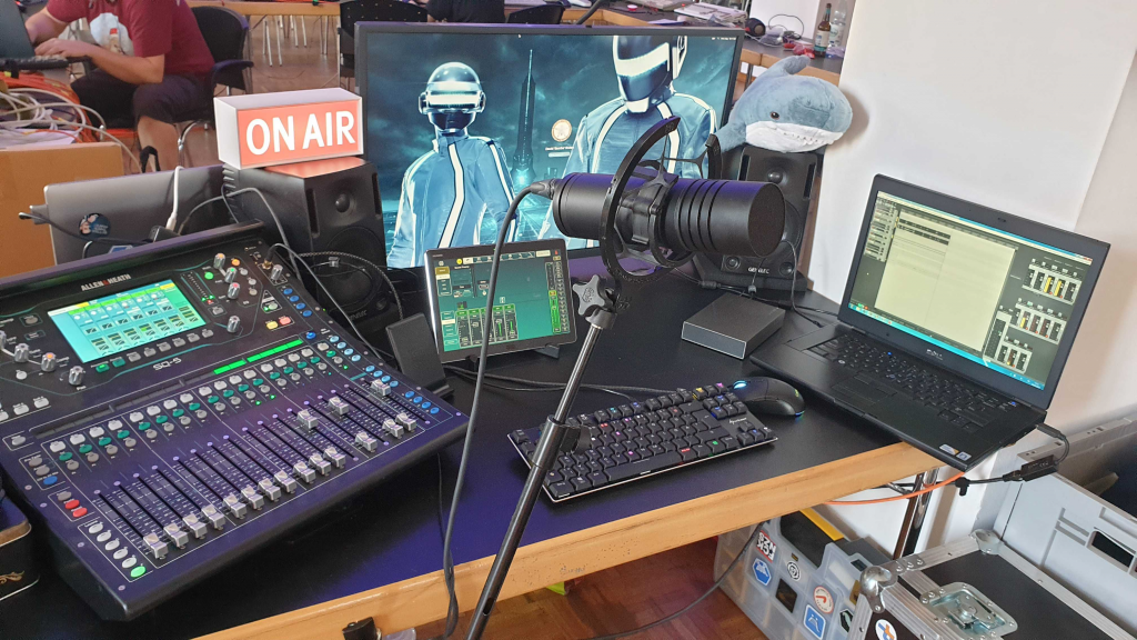 A mixing desk, a microphone, a large screen, a tablet running remote control software, a laptop running recording software, external harddisk, keyboard, mouse, and BLÅHAJ overseeing everything.